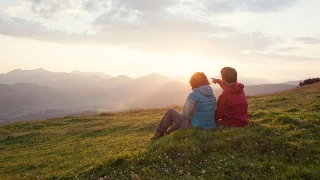 Ein Paar sitzt waehrend einer Wanderung auf der Wiese und blickt ueber das Tal.